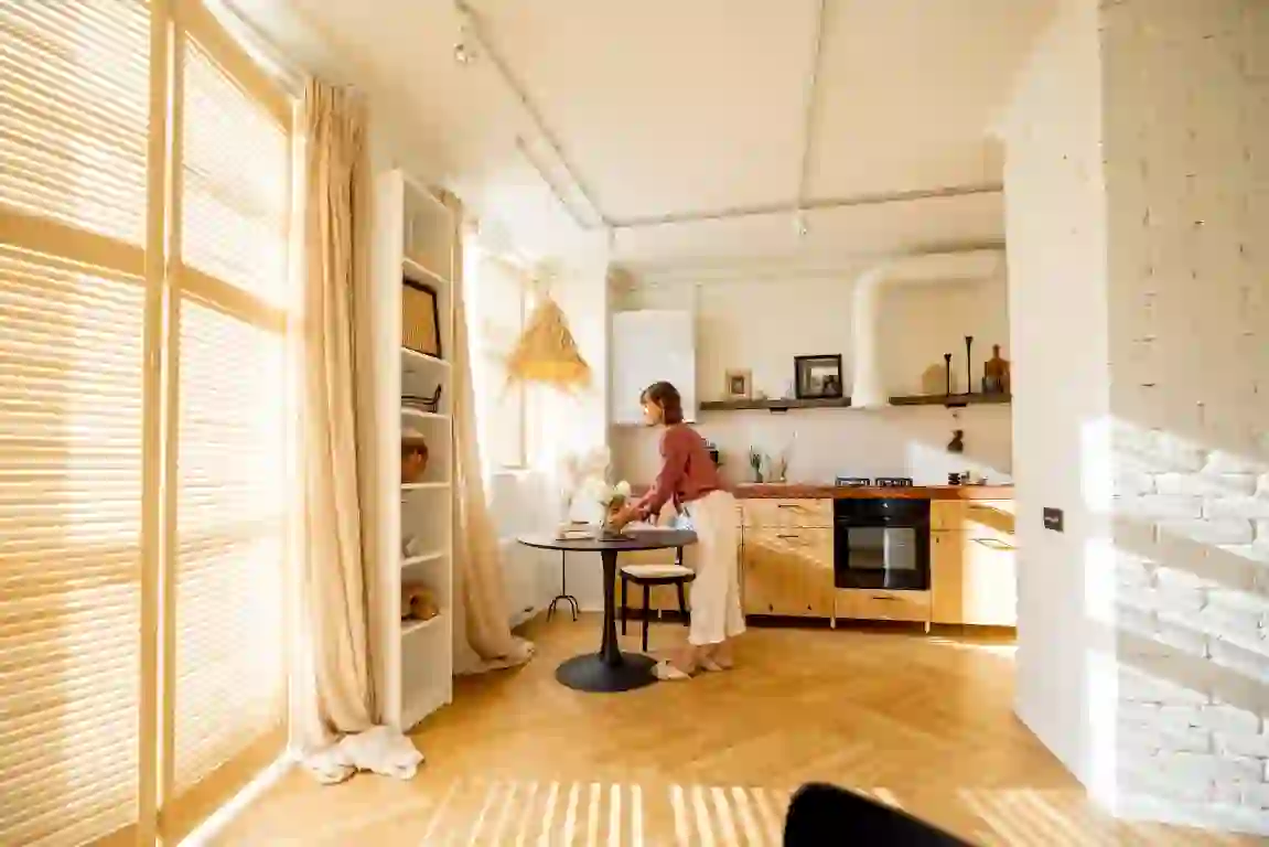 A woman stands at a small round table in a bright Lakeland kitchen with wooden cabinets, white walls, and large windows covered by sheer curtains. The room is warmly lit, with natural light streaming in, highlighting the hardwood floor and modern decor.
