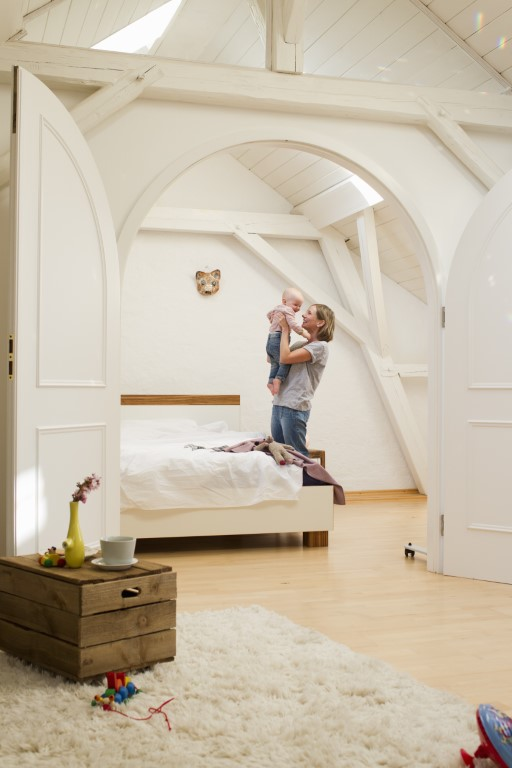A woman cradles a baby in a bright, airy bedroom with white walls and wooden beams. An arched door leads to the room, giving it an air of home mystery. A wooden crate with a cup and flower rests on the floor, while a cozy rug and scattered toys complete the scene.