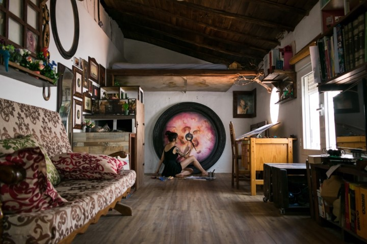 A cozy attic studio with wooden floors and beams exudes functionality. A woman sits by a large, circular painting, working intently. The room's framed artwork, bookshelves, and a patterned sofa create an artistic and inviting atmosphere tinged with mystery.