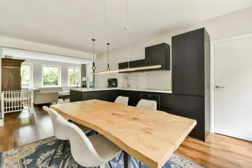 Modern kitchen and dining area renovation in Lakeland, Florida featuring a light wooden table with four white chairs, black cabinets, pendant lighting, hardwood floors, and a patterned rug. Living room and large windows in the background make this layout suitable for kitchen cabinet installation, home additions, or mother-in-law suite projects.