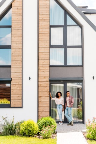 A couple stands together outside the entrance of a modern house with large windows, wooden accents, and a well-kept garden—a true showcase of valuable renovations inspiring Florida homeowners.