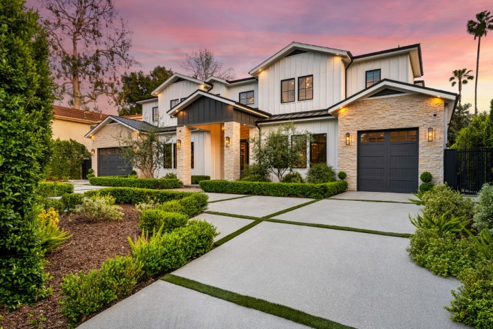 Modern two-story house with stone and white siding exterior, dark garage doors, manicured landscaping, and a sunset sky in the background. Ideal for aging in place renovations, the concrete driveway features green grass strips between slabs.