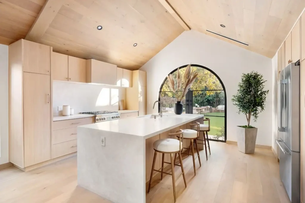 Modern kitchen renovation in Lakeland, Florida with light wood cabinets and flooring, a large white island with three bar stools, and a tall indoor plant. Sunlight from an arched window overlooking a green backyard highlights features ideal for kitchen cabinet installation, home additions, or mother-in-law suite designs.