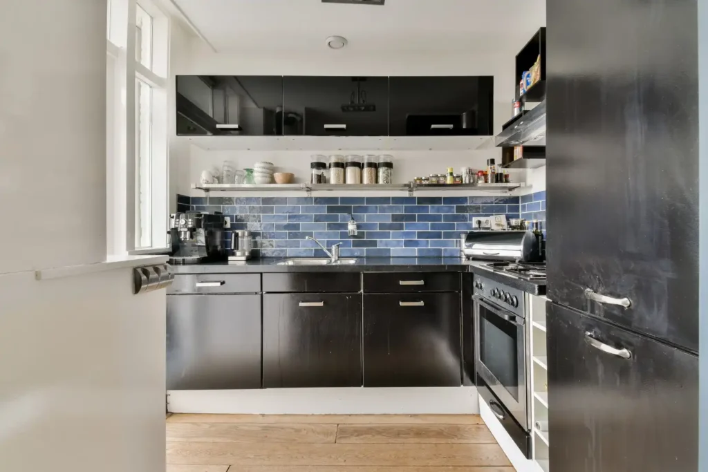Modern kitchen renovation in Lakeland, Florida featuring black cabinets, blue subway tile backsplash, open shelving with jars and dishes, stainless steel appliances, and wooden flooring. Natural light from a side window enhances this space, ideal for kitchen cabinet installation, home additions, or mother-in-law suite layouts.