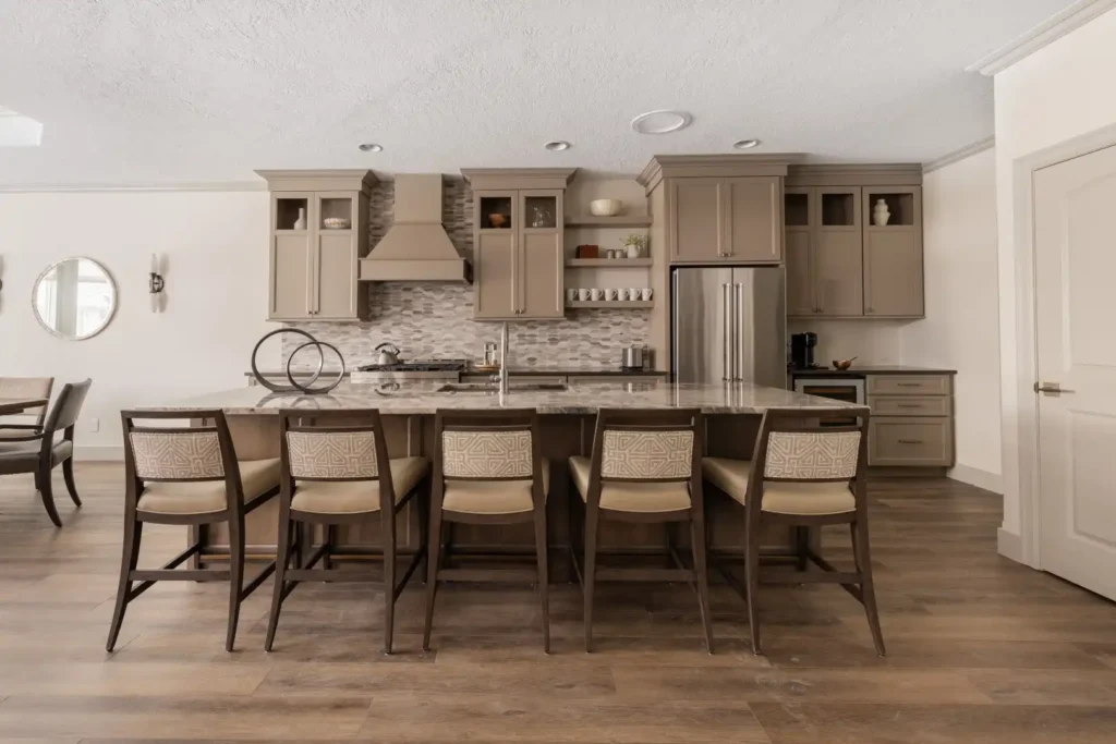 Modern kitchen renovation in Lakeland, Florida with tan cabinets, stainless steel appliances, tile backsplash, and a large island with seven cushioned bar stools. Features include wood flooring, neutral decor, and a visible dining area, ideal for home additions, kitchen cabinet installation, or mother-in-law suite upgrades.