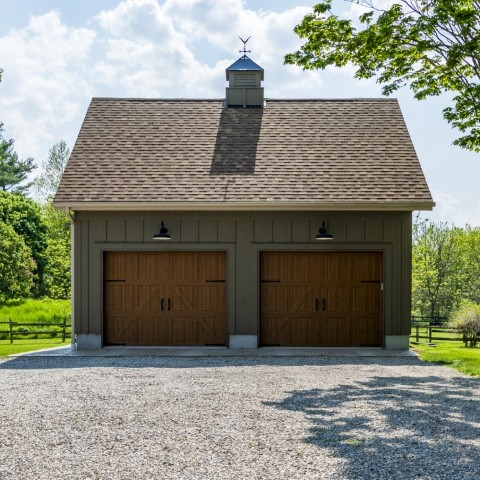 A detached garage with brown wooden doors, gray siding, a shingled roof, and a small cupola on top sits amid trees and greenery under a partly cloudy sky.