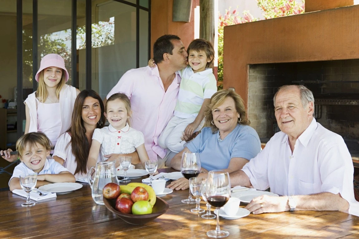 Three generations of a family, including children, adults, and grandparents, sit together at a table outdoors, smiling—reflecting the warmth and joy that thoughtful multi-generational home design brings to family gatherings.