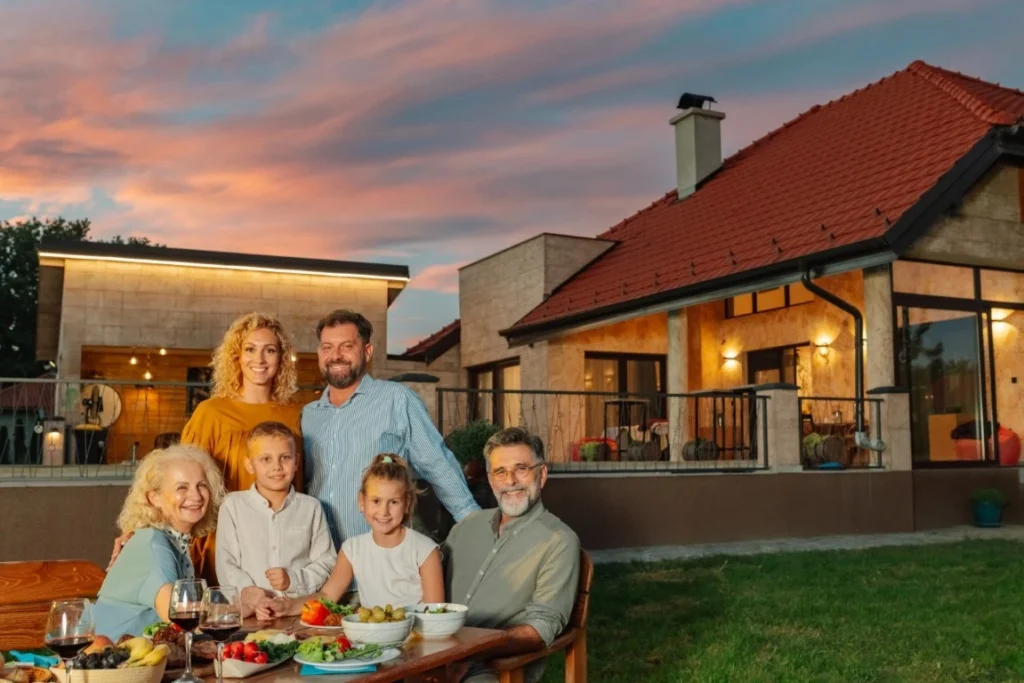 Three adults and three children sit and stand around a table with food, smiling in front of a modern house at sunset. The colorful sky and cheerful atmosphere highlight the joy of multi-generational home design.