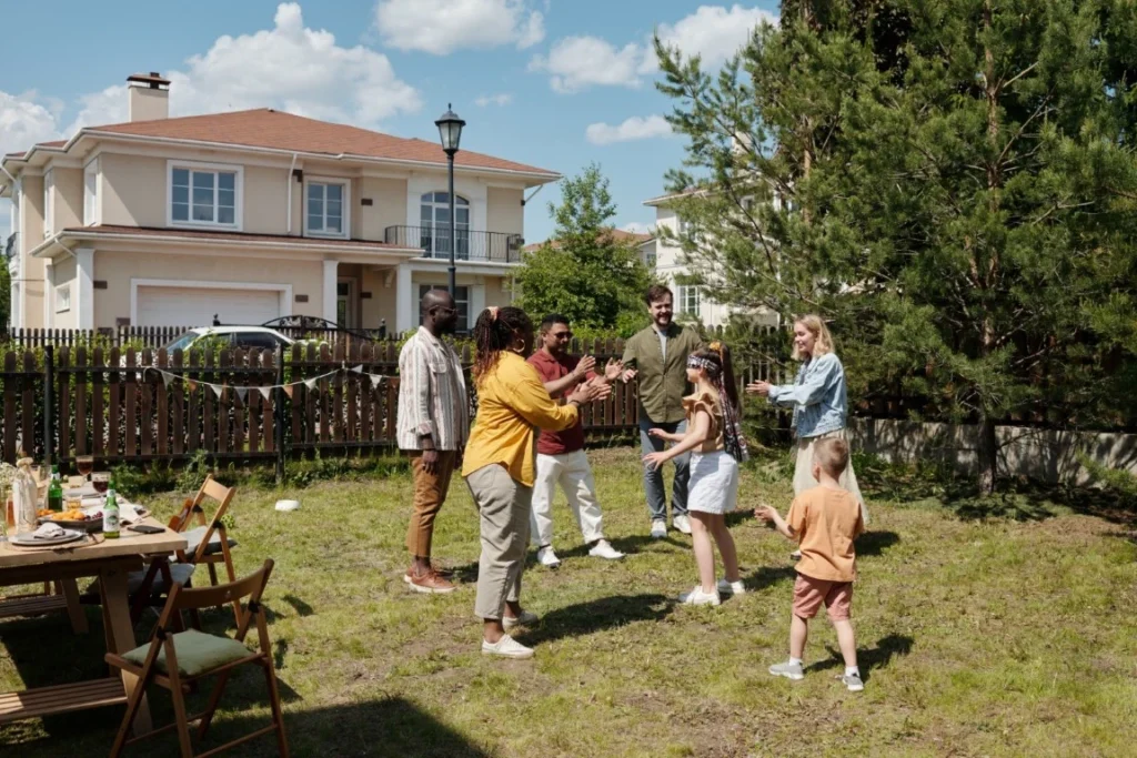 A group of adults and children play together in a backyard on a sunny day, enjoying a casual outdoor gathering that reflects the spirit of multi-generational home design, with a house, trees, and dining table in the background.