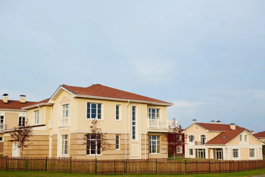 A large, two-story yellow house with white trim and a red-tiled roof showcases multi-generational home design, standing in front of a wooden fence, with a similar house in the background under a clear sky.