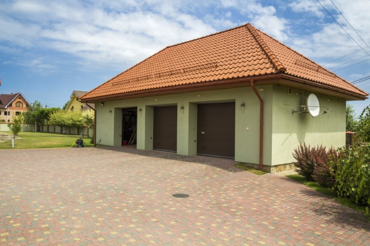A detached garage with a red tile roof, light green walls, and two closed brown garage doors stands in a grassy yard. Featuring a satellite dish, paved driveway, and surrounded by bushes beneath a partly cloudy sky.