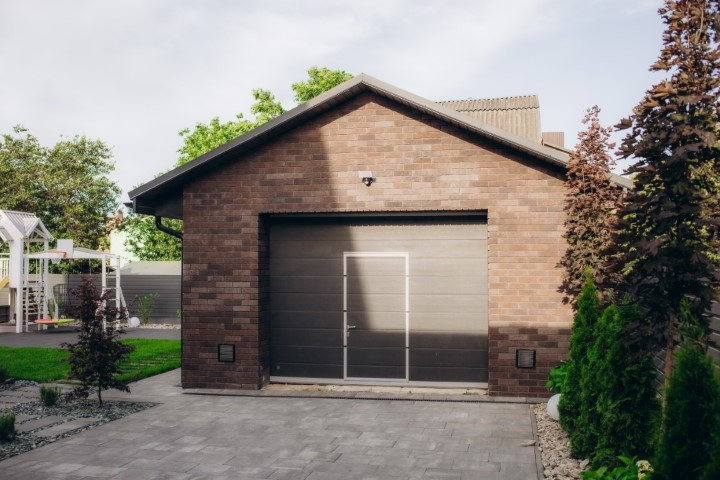 A modern detached garage with brown brick walls and a closed gray door, surrounded by a tidy yard featuring green grass, shrubs, and a paved driveway—perfect for anyone seeking the convenience of a spacious detached garage.