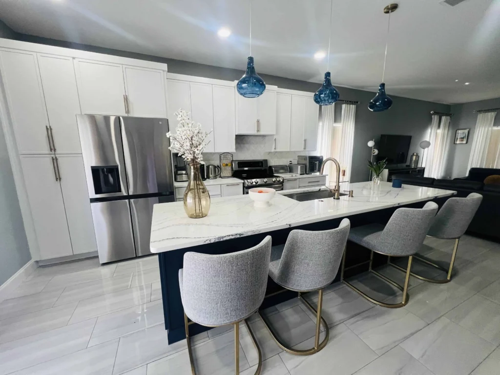 Modern Lakeland kitchen with white cabinets, stainless steel appliances, a marble island with four gray barstools, blue pendant lights, and decorative vases—perfect for stylish FL living.