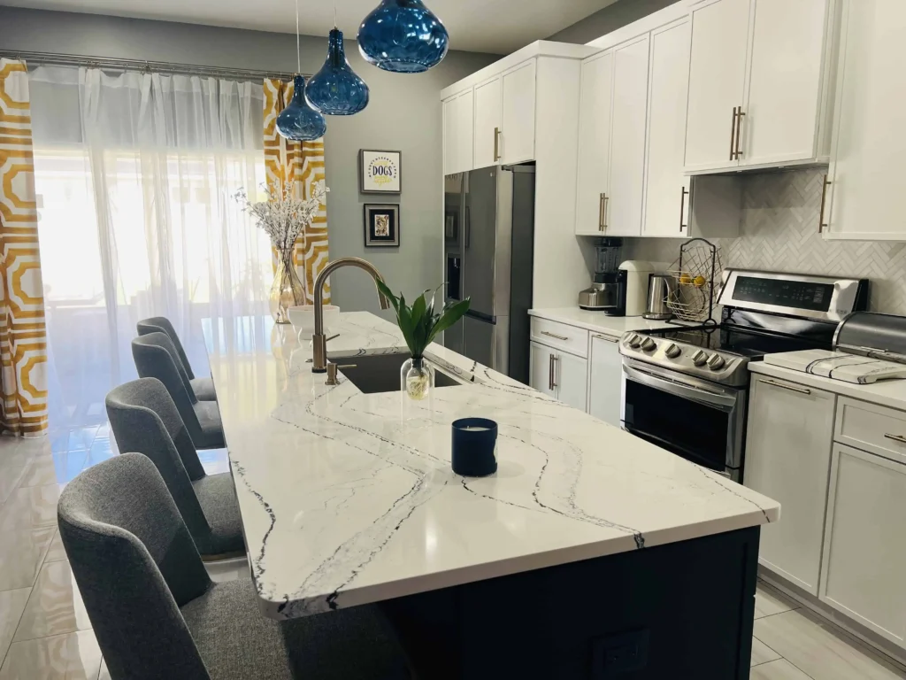Modern kitchen in Lakeland, FL with white cabinets, stainless steel appliances, marble island with bar stools, blue pendant lights, and yellow patterned curtains by a large window.