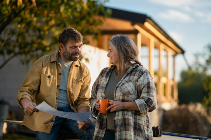 A man and woman talk outdoors near a house under construction in Lakeland, FL; the man holds a blueprint, and the woman holds an orange mug.