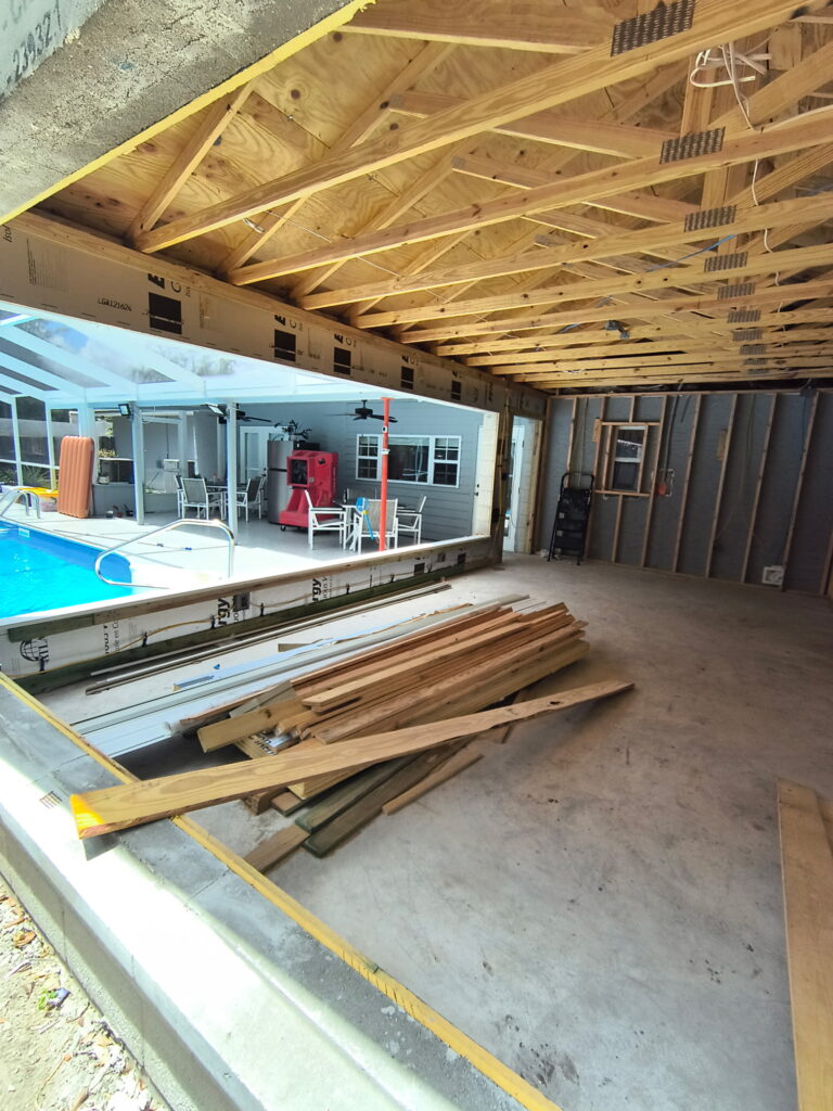 View of a partially constructed garage in Lakeland, FL, with exposed wooden framing and ceiling, construction materials on the floor, and a pool area visible through an open wall.