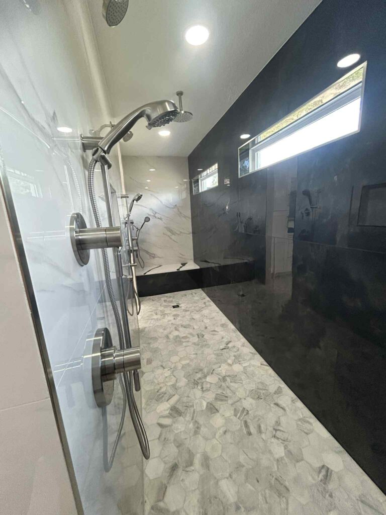Modern walk-in shower in Lakeland, FL, featuring dual stainless steel showerheads, marble hexagon tile floor, black and white marble walls, and a horizontal window letting in natural light.