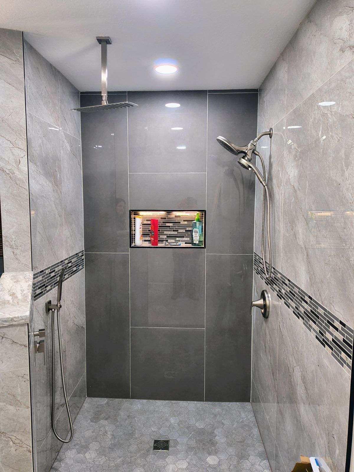 Modern walk-in shower in Lakeland, FL, with gray tiles, a rainfall showerhead on the ceiling, handheld shower on the wall, and a built-in niche holding toiletries.
